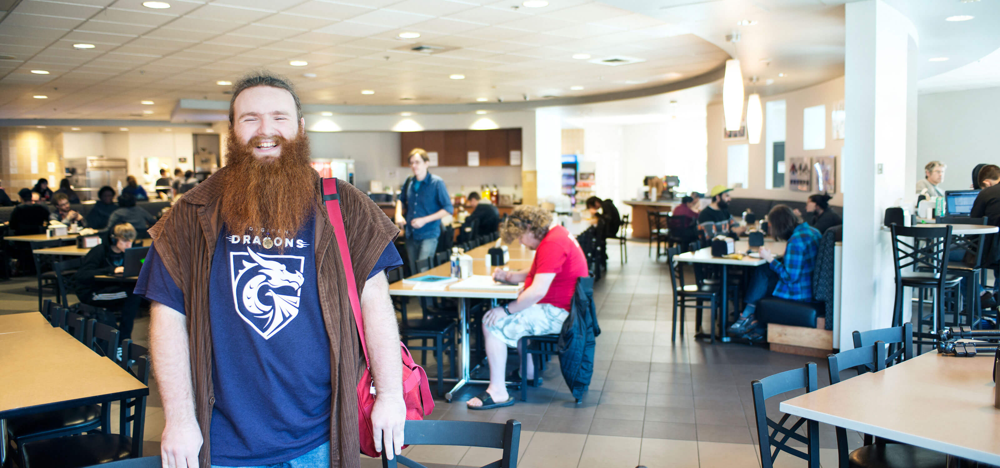 A smiling, bearded man wearing a purple, dragon-logo t-shirt stands in a dining hall where others are seated at tables.