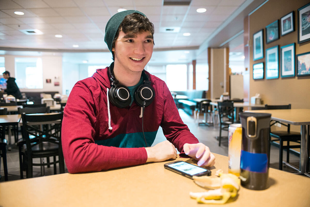 A smiling man in a maroon parka with headphones draped over his neck sits at a table in a brightly lit dining hall.