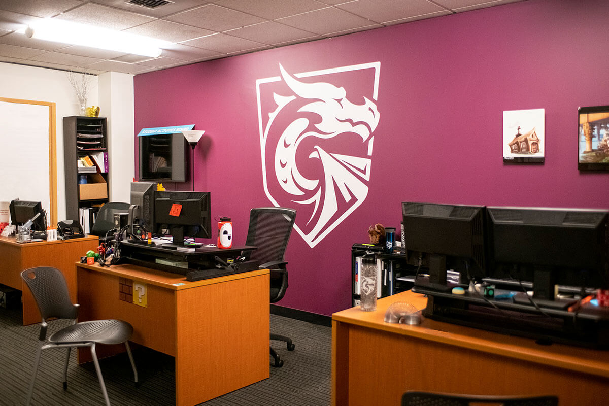 An office space with desks and computers in front of a maroon-colored wall with a large white logo of a dragon painted on it.