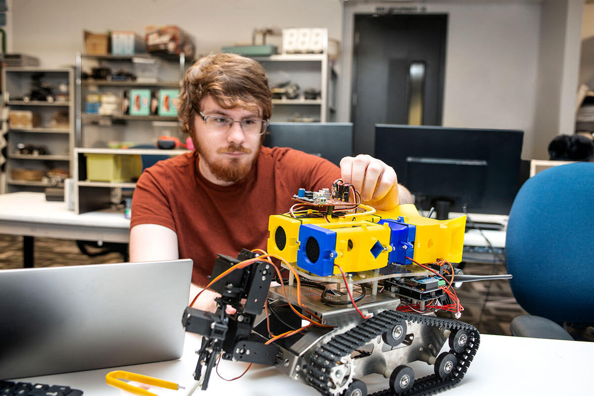 A man in an orange t-shirt sits working on a blue-and-yellow robot with exposed circuits, plastic, metal, and treads.