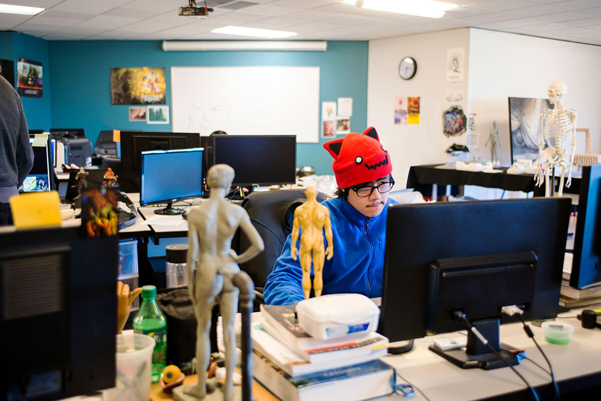 A man in a blue parka and red beanie sits at a computer in a room filled with anatomical models, both miniature and life-size