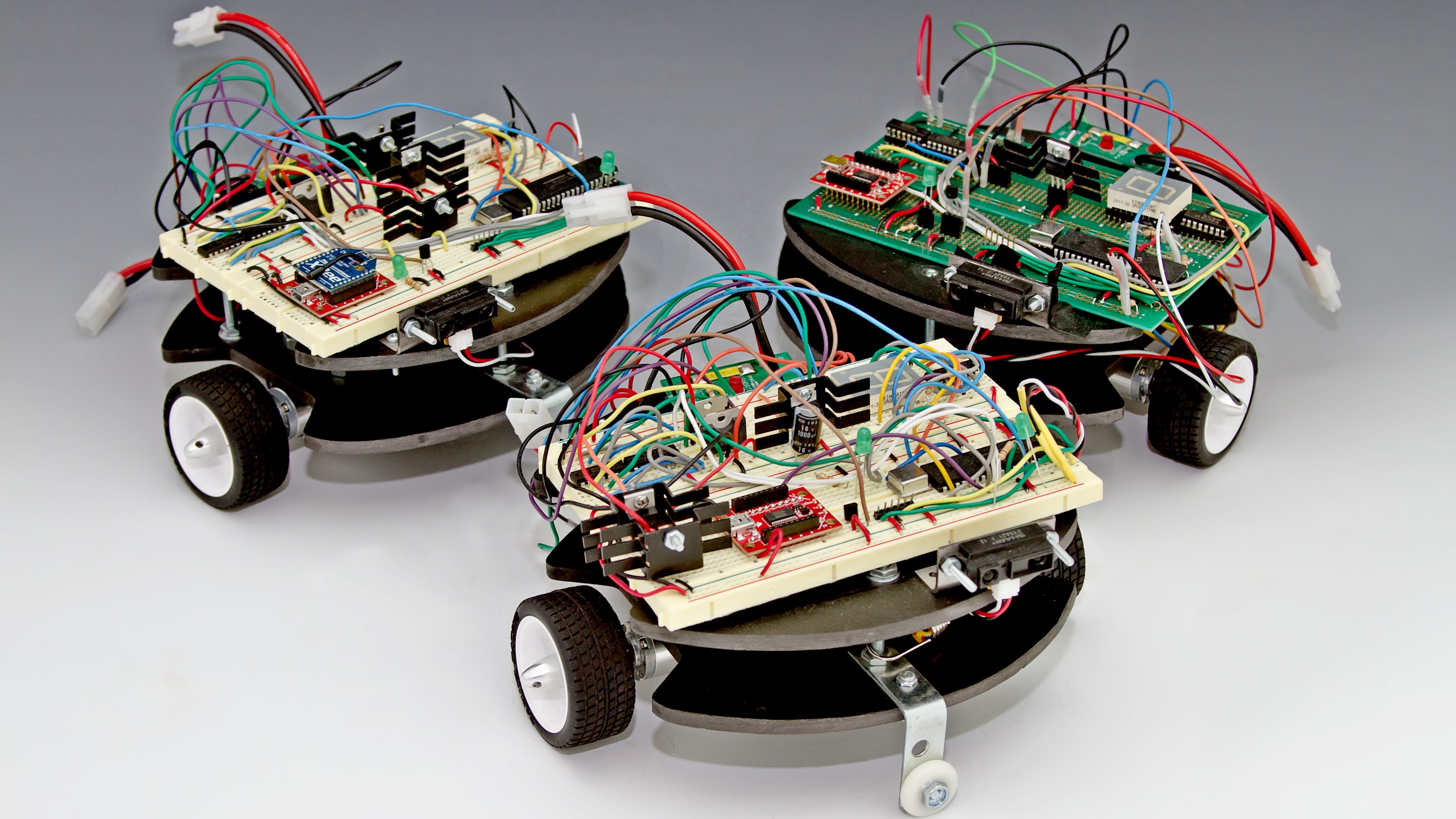 Three square robots with rounded fronts, black and white wheels and exposed circuitry.