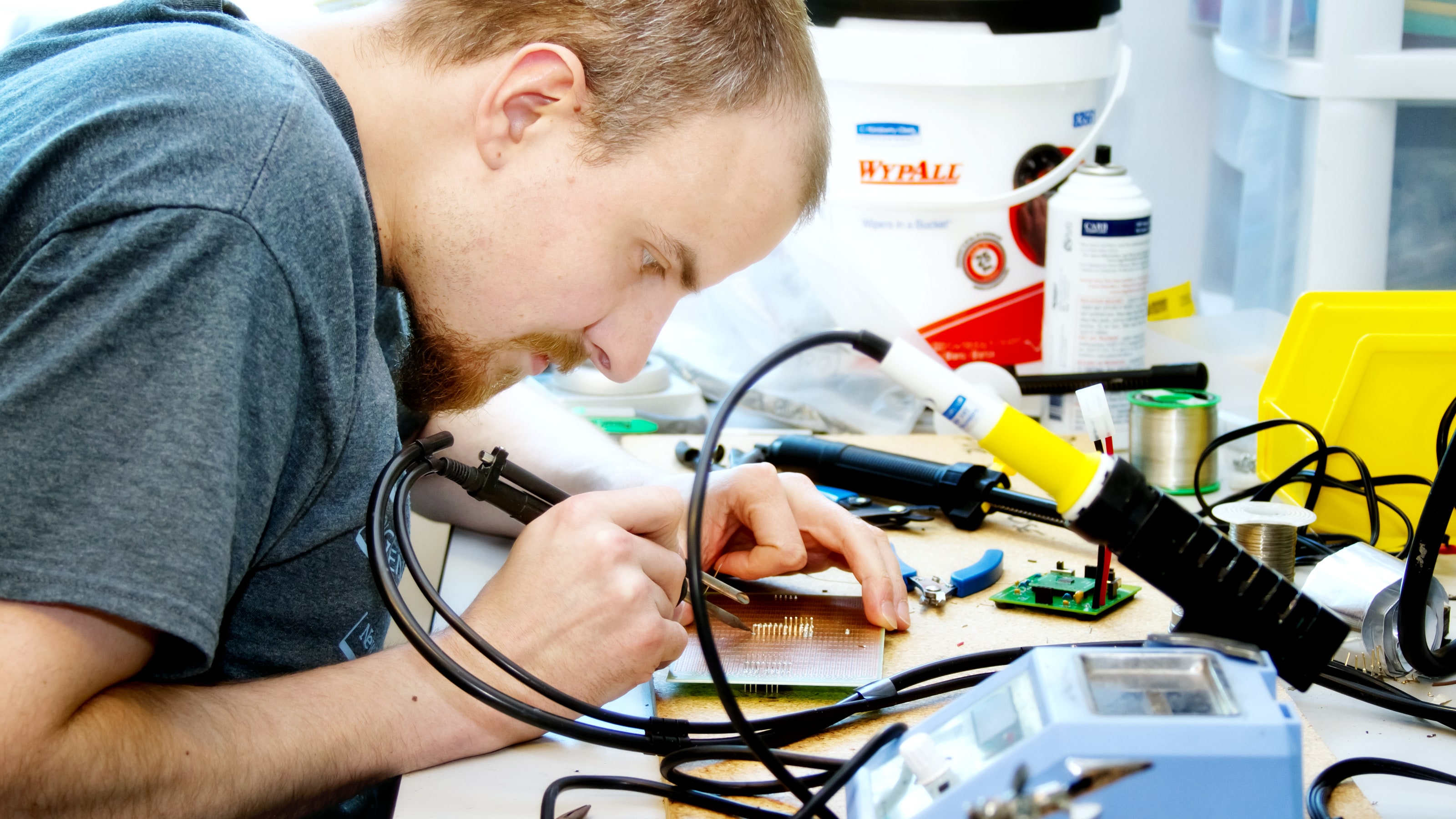 A student solders a circuit board on a desk cluttered with tools.