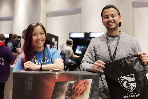 Two employees at a convention hall pose with a black DigiPen Dragons drawstring backpack and boxed computer monitor.