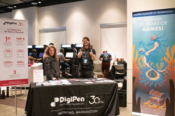 Two employees stand behind a DigiPen convention table in front of a row of arcade cabinets.