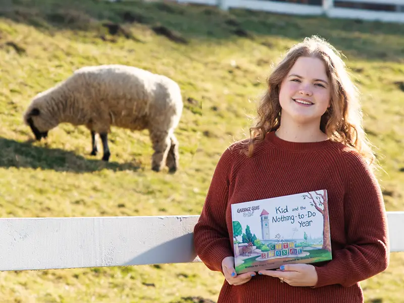 Melissa Murakami holds her kid’s book in front of a field with sheep in it.