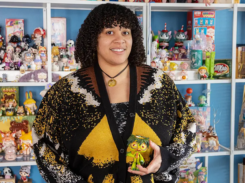 Nena Ijiomah smiles in front of a row full of toy figurines, holding a small green kappa figurine in her hand.