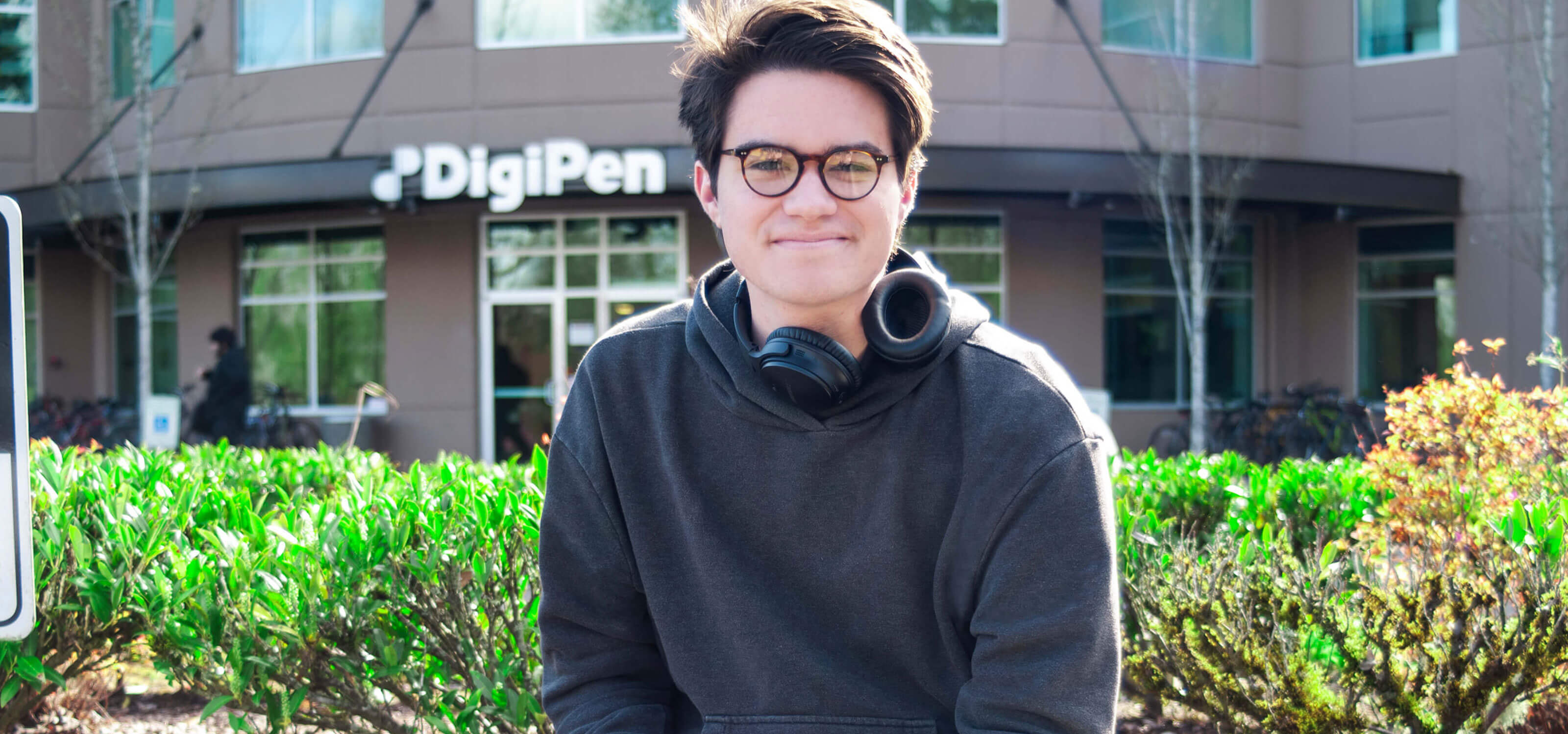 A student wearing headphones smiles on a sunny day in front of the DigiPen building