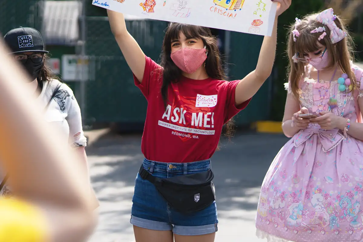 A DigiPen voulunteer smiling outdoors holding a large colorfully decorated sign.