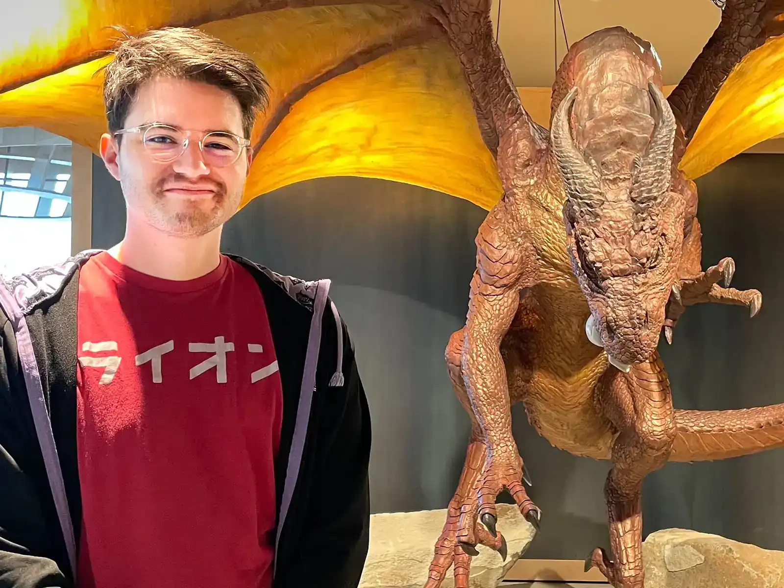 PJ Rivas smiles in front of a large lifelike dragon statue in the Wizards of the Coast office building. 