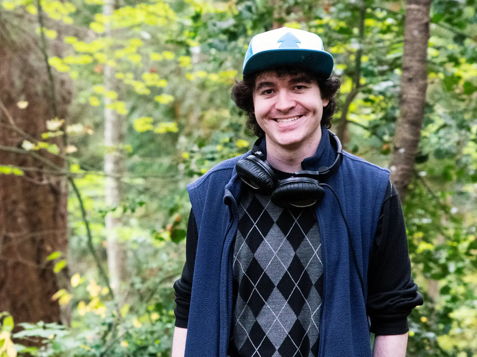 Chris Onorati smiles in the woods next to a tree wearing his iconic Gravity Falls hat and vest.