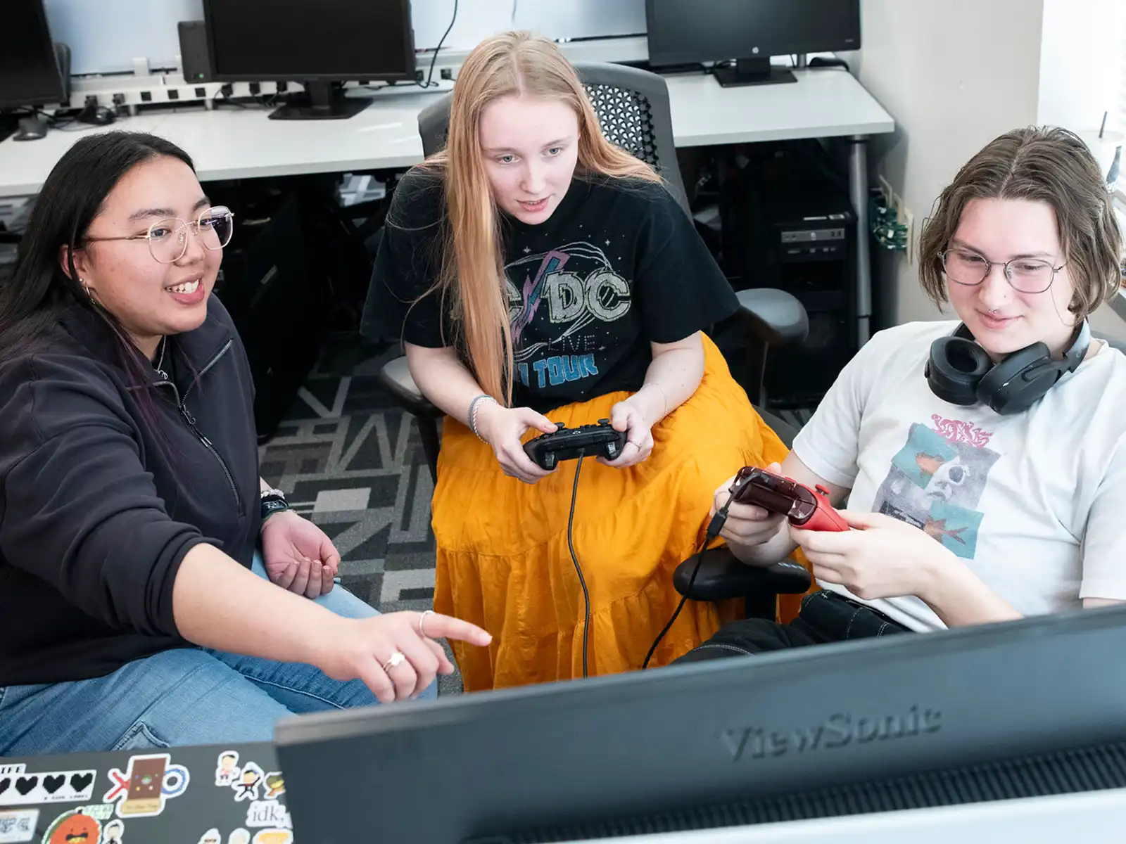 Two DigiPen students at Playfest hold controllers and play a computer game as a third student points to the screen.