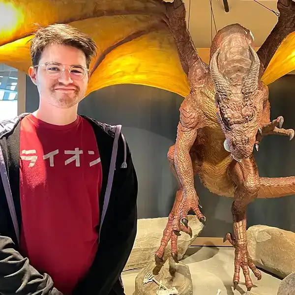 PJ Rivas smiles in front of a large lifelike dragon statue in the Wizards of the Coast office building. 