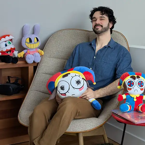 Evan Alderete poses in a chair holding a Pomni plushie surrounded by other stuffed dolls of Amazing Digital Circus characters.