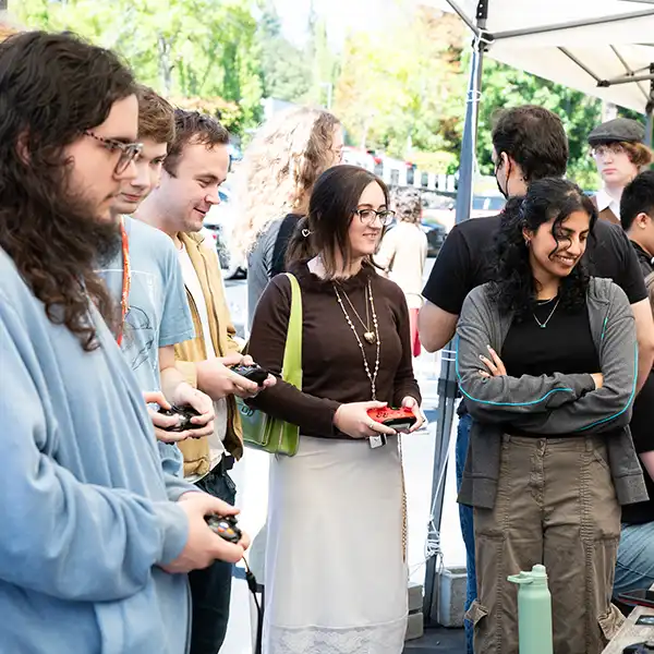 Four DigiPen students holding controllers play a game side-by-side with one onlooker standing outside of campus.