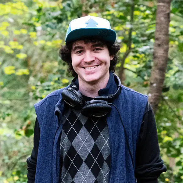 Chris Onorati smiles in the woods next to a tree wearing his iconic Gravity Falls hat and vest.