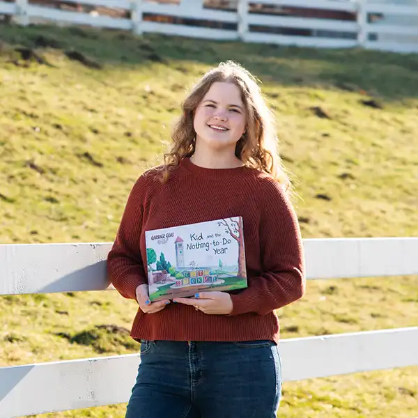 Melissa Murakami holds her kid’s book in front of a field with sheep in it.