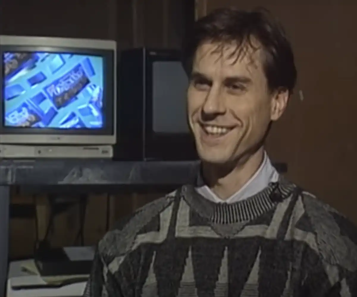 Matt Brunner from 1994 smiles at the camera in sweater sitting in front of a TV displaying a Chips Ahoy commercial.