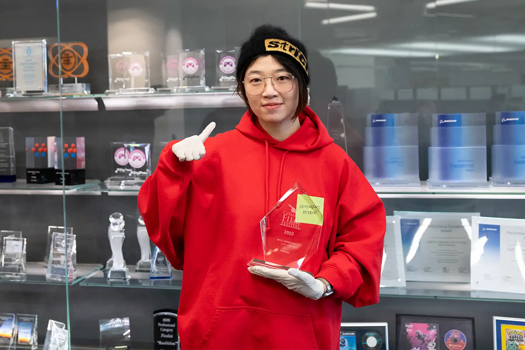 Mingyuan Li holds a clear trophy and makes a number one hand sign in front of DigiPen’s campus trophy case.