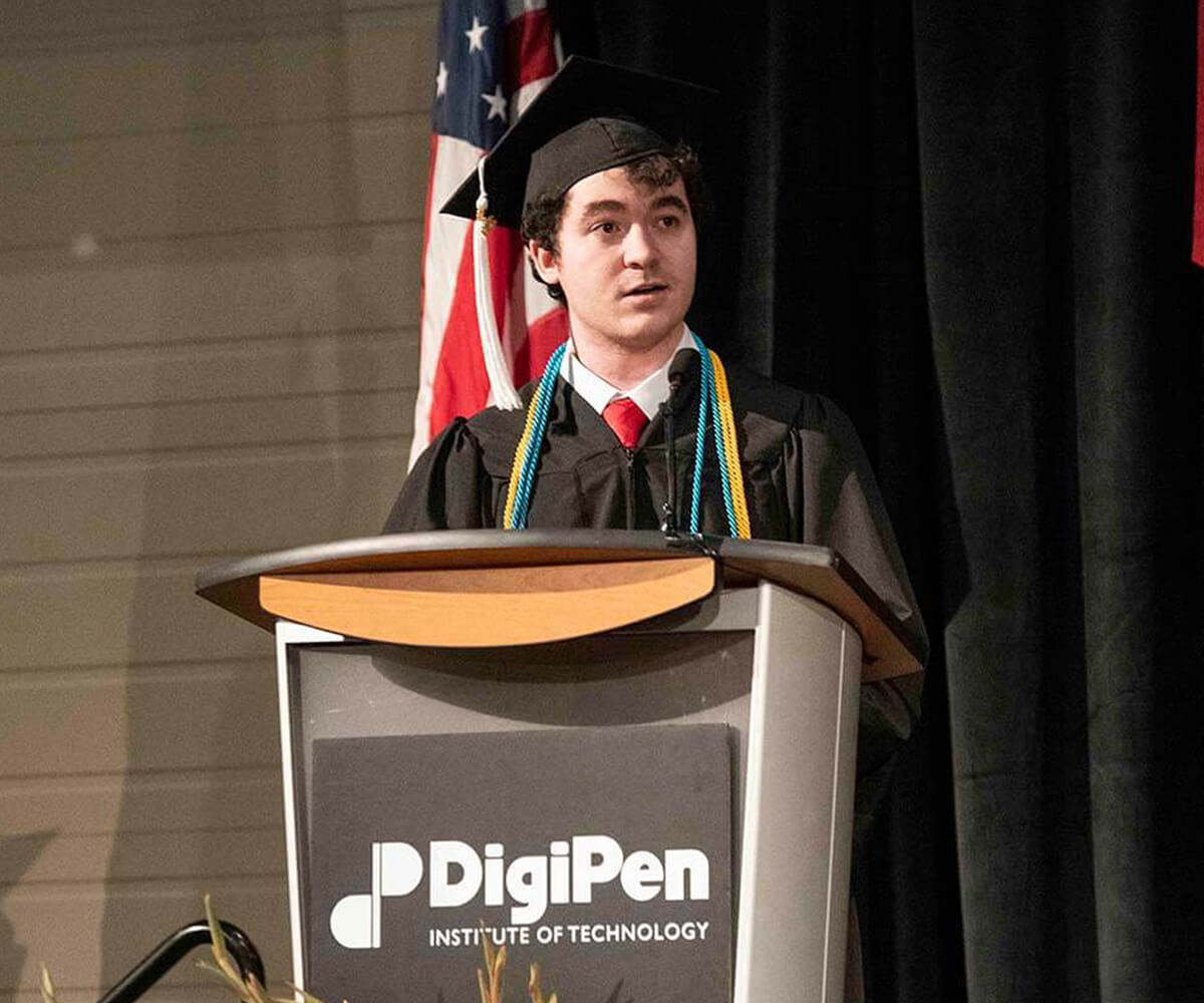 Chris Onorati speaks wearing a graduation robe and hat at a podium with the DigiPen logo on it.